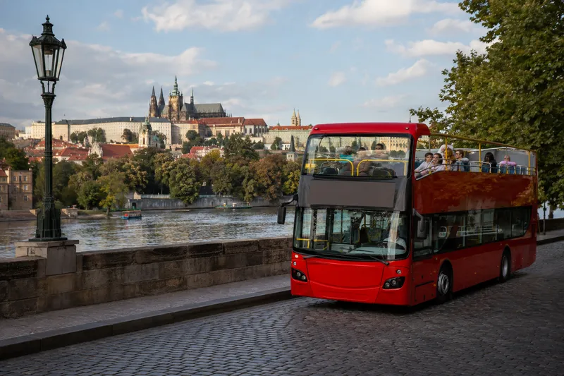 Red double-decker sightseeing bus passing Prague Castle along the Vltava River